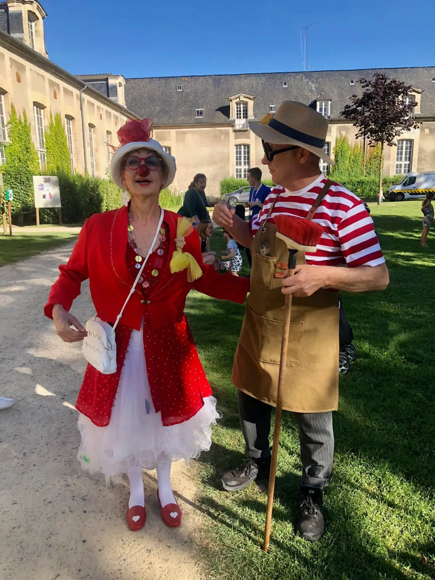 Deux clowns en déambulation dans un parc : l'une en veste et robe rouges à pois, coiffée d'un chapeau fleuri ; l'autre en marinière rayée et tablier de travail, un balai à la main.