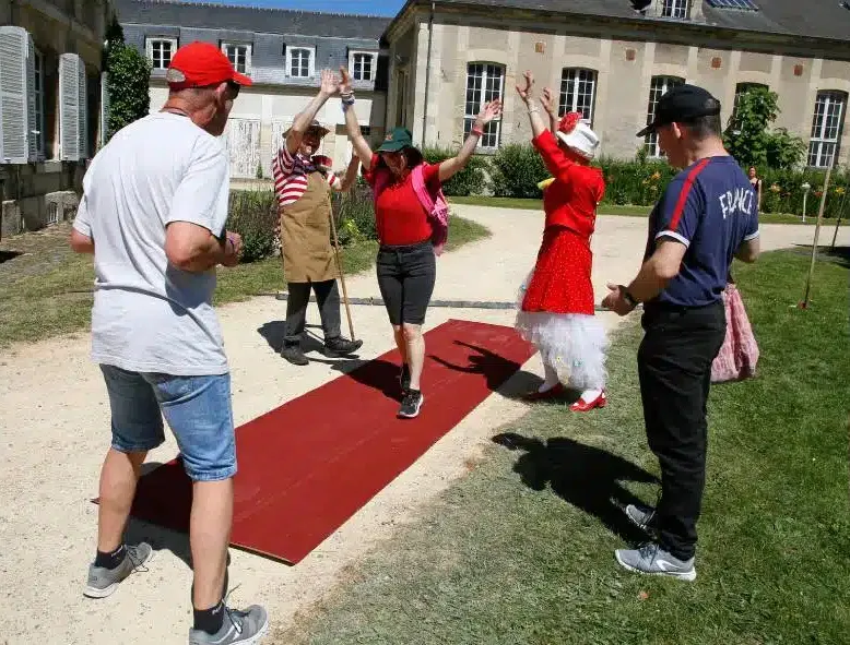 Les clowns déroulent un tapis rouge sur une allée et lèvent les bras pour accueillir une passante, sous le regard amusé de deux autres personnes.